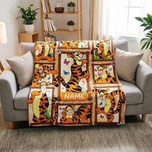 May include: A tan fleece blanket featuring multiple rectangular frames containing illustrations of the orange and black striped character Tigger. The design includes colourful butterflies and a central orange banner with the text NAME.