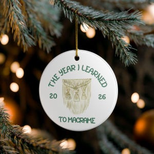 May include: A round white ceramic ornament featuring a green printed illustration of a macrame wall hanging. The text reads THE YEAR I LEARNED TO MACRAME with the year 2026 split on either side of the graphic. A gold string is attached to the top.