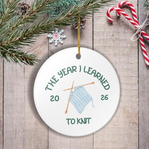 Può includere: Un ornamento rotondo in ceramica bianca con una grafica di tessuto a maglia azzurro su due ferri da maglia in legno. Il testo THE YEAR I LEARNED TO KNIT è stampato con l'anno 2026 su entrambi i lati. Un cordino dorato è fissato in alto.