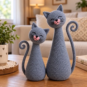 Pode incluir: Duas figuras de gato em crochê cinza de alturas diferentes, cada uma com orelhas pontudas, um pequeno nariz costurado em rosa e uma boca bordada em preto com olhos fechados. Ambos os gatos têm corpos esguios e caudas enroladas.