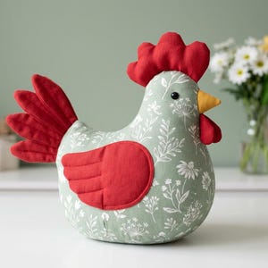 May include: A stuffed fabric chicken toy featuring a sage green body with a white botanical floral print. The chicken has a bright red comb, tail feathers, and a wing, along with a yellow beak and a small black bead eye for a detailed appearance.