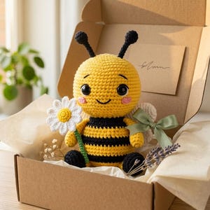 May include: A crocheted bee plush toy with a yellow and black striped body, black antennae, and large dark eyes. The bee holds a white crocheted daisy with a yellow centre. It is nestled inside a brown cardboard gift box with tissue paper.