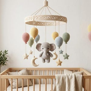 May include: A crochet baby mobile featuring a central grey elephant surrounded by hanging pastel-coloured balloons, stars, and crescent moons. The mobile is suspended from a circular, cream-coloured, textured frame with intricate woven patterns.