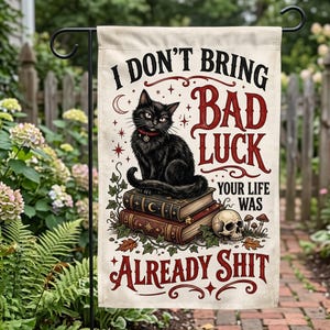 Könnte beinhalten: Eine rechteckige Gartenflagge mit einem schwarzen Katzenmotiv, die auf zwei gestapelten Büchern sitzt, sowie einem menschlichen Schädel und Pilzen. Der Text lautet: I DON'T BRING BAD LUCK YOUR LIFE WAS ALREADY SHIT. Das Design enthält Sterne, eine Mondsichel und Efeublätter.