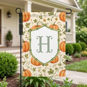 May include: A rectangular garden flag featuring a repeating pattern of orange pumpkins and green vines on a cream background. In the centre, a white shield-shaped emblem framed by green bamboo-style stalks displays a large, sage green letter H.