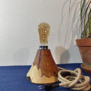 Pode incluir: Um candeeiro de mesa com base cónica em madeira natural com veios visíveis e bordas de casca. O candeeiro é rematado com uma lâmpada de vidro transparente estilo Edison com um filamento laranja em espiral e um cabo de tecido bege.