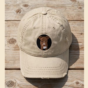 Puede incluir: Una gorra de béisbol beige con un acabado desgastado. La parte frontal presenta un parche circular negro bordado con la ilustración de un perro marrón. El perro tiene una mancha blanca en la frente y la barbilla, centrada en el panel frontal.
