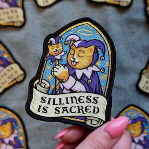 May include: Embroidered patch featuring a cartoon cat in a jester's outfit, holding a staff with a cat head. The patch has a banner that reads "Silliness is Sacred". The cat is orange and white, with a purple and blue background.