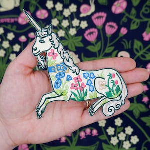 Puede incluir: Parche blanco bordado de un unicornio con detalles florales en tonos rosa, azul y verde. El unicornio lleva un collar azul y dorado.
