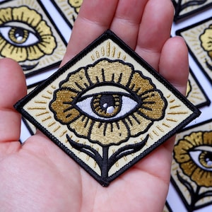May include: A gold embroidered patch with a stylized eye inside a flower. The eye is white with a brown iris and black pupil. The flower is gold with a black outline and a green stem. The patch is on a white background.