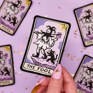 May include: A tarot card patch featuring "The Fool" with a jester figure, black cat, and text. The patch has a black border, a purple background, and a yellow sun. The Fool is depicted in white and black, carrying a bag and staff.