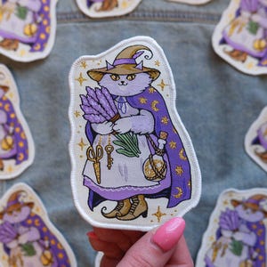 May include: A whimsical patch featuring a lavender-colored cat dressed as a witch, wearing a pointed hat and cape. The cat holds lavender and a potion bottle. Gold accents and stars decorate the design. The patch is approximately 3 inches tall.