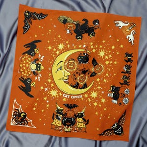May include: Orange Halloween-themed bandana with a whimsical design. The central image features a smiling moon with a cat in a witch hat. Surrounding the moon are stars, bats, ghosts, and other Halloween-related illustrations. The text "CAT COVEN" is also visible.