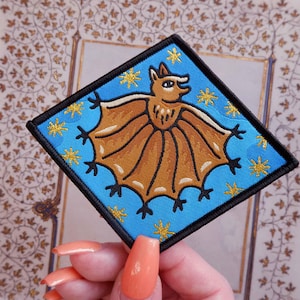 May include: Diamond-shaped embroidered patch with a brown bat design on a light blue background. The bat has outstretched wings and is surrounded by gold star motifs. The patch has a black border, suitable for clothing or accessories.