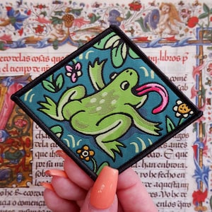 May include: A square patch featuring a green frog with a pink tongue, set against a teal background with floral accents. The patch has a black border and is likely an iron-on or sew-on applique.