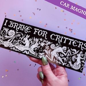 Peut inclure: Aimant de voiture noir avec texte et illustrations blancs. L'aimant indique "I Brake for Critters" et présente des dessins détaillés de lapins, de squelettes et d'autres créatures. Une bannière violette en haut indique "CAR MAGNET!"