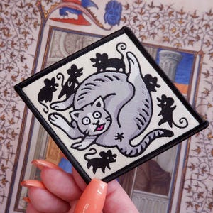 May include: Square patch featuring a cartoon cat surrounded by black mice. The cat is gray with a pink tongue and wide eyes. The patch has a black border and is on a cream-colored background.