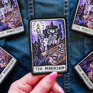 Peut inclure: Un écusson brodé rectangulaire représentant une carte de tarot intitulée THE MAGICIAN. L'illustration montre une silhouette portant un chapeau et tenant une bougie allumée, entourée de chats noirs, avec une épée et un pentacle sur une table.