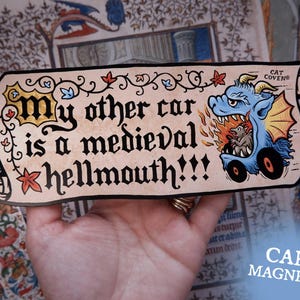 Op de afbeelding: Een rechthoekige auto magneet met de tekst "My other car is a medieval hellmouth!!!" in een middeleeuws lettertype. Een blauwe draak met vlammen en een klein wezen staat rechts. De magneet wordt in een hand gehouden.