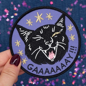 May include: Purple embroidered iron-on patch with a black cat with a wide open mouth and a pink tongue. The cat is surrounded by gold stars and the text "GAAAAAY!!!" in a purple font.