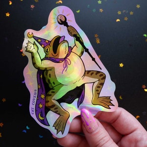 May include: A holographic sticker featuring a cartoon frog wearing a purple wizard hat and robe, holding a wand. The frog is green with brown spots and has a large, toothy grin. The sticker has a rainbow iridescent finish and says "CAT COVEN®" in purple text.