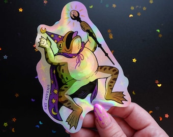 May include: A holographic sticker featuring a cartoon frog wearing a purple wizard hat and robe, holding a wand. The frog is green with brown spots and has a large, toothy grin. The sticker has a rainbow iridescent finish and says "CAT COVEN®" in purple text.