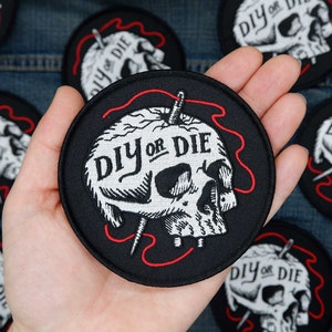 Puede incluir: Un parche bordado negro con una calavera blanca y el texto "DIY or DIE" en una fuente estilizada. La calavera está cosida con hilo rojo.