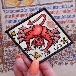 May include: Square patch featuring a red devil with bat wings, a long tail, and a downturned face. The patch has a black border and is on a cream-colored background. Gold star and floral accents are in the corners. The background is an old manuscript.