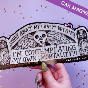 Könnte beinhalten: Ein Auto-Magnet mit einem Grabstein-Design. Der Magnet zeigt einen Schädel mit Flügeln und den Text "Sorry about my crappy driving! I'm contemplating my own mortality!!!" Die Website "CATCOVEN.com" ist ebenfalls sichtbar.