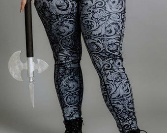 Puede incluir: Leggings negros con un patrón floral blanco y gris con un diseño repetitivo de hojas y flores estilizadas. Los leggings son usados por una persona que sostiene un hacha plateada.