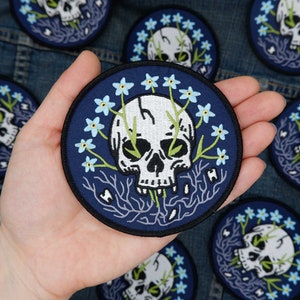 May include: A blue embroidered patch featuring a white skull surrounded by blue forget-me-not flowers. The skull has green eyes and is set against a dark blue background.
