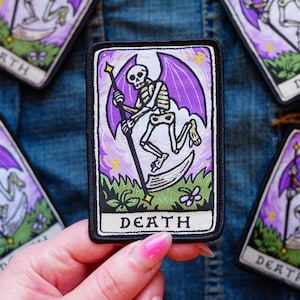 Peut inclure: Un écusson brodé rectangulaire représentant un squelette aux ailes violettes tenant une faux, sur un fond de ciel violet et d'herbe verte. Le mot DEATH est inscrit en lettres capitales noires sur une bannière blanche en bas de l'écusson.