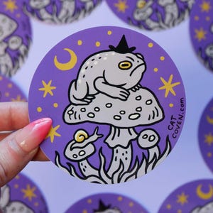 Op de afbeelding: Paarse sticker met een cartoon kikker die een heksenhoed draagt ​​en op een paddenstoel zit, met een slak ernaast. De achtergrond is een paarse nachthemel met sterren en een halve maan. De tekst op de sticker is "CAT COVEN.COM".