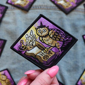 May include: Square patch featuring a cartoon owl wearing a witch hat, quill, and open book with candles and a potion bottle. The patch has a black border and a purple and gold color scheme. The patch is on a denim background.