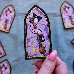 May include: Embroidered patch featuring a whimsical witch riding a broom against a purple sky with a crescent moon and stars. The patch has a black border with gold accents. The witch wears a black hat and boots. The patch is held by a person with pink nail polish.