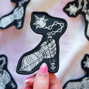 May include: A black embroidered patch featuring a silver metallic thread design of a gauntleted hand holding a spiked flail weapon. The patch has a black stitched border and a detailed illustration of armor plating on the hand and wrist.