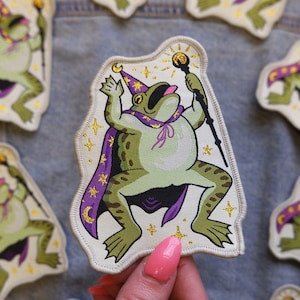 May include: A whimsical patch featuring a green frog dressed as a wizard, wearing a purple cape and hat with a crescent moon. The frog holds a staff with a golden orb. Gold stars and accents surround the frog.