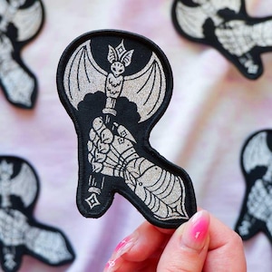 May include: A black embroidered patch featuring a silver metallic thread design of a gauntleted hand gripping the handle of a battle axe. The axe head is shaped like a bat with spread wings. The patch has a black stitched border edge.