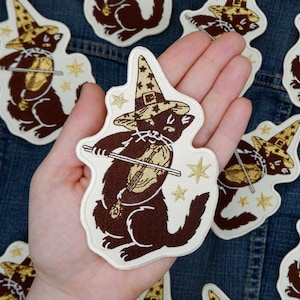 May include: A hand holding a brown and white embroidered patch of a black cat wearing a wizard hat and playing a violin. The cat is surrounded by small gold stars.