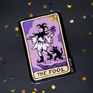 Könnte beinhalten: Ein Tarotkarten-Aufkleber mit der Karte "Der Narr". Die Illustration zeigt eine Narrenfigur mit einer schwarzen Katze vor einem lila Hintergrund. Die Karte hat einen schwarzen Rand und der Text "CAT COVEN" ist auf der Seite aufgedruckt.