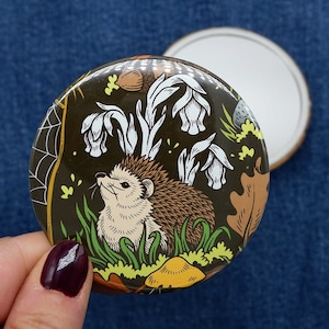 May include: A round, compact mirror with a black background and a whimsical illustration of a hedgehog surrounded by white flowers and green leaves. The hedgehog is brown and white with a pink nose.