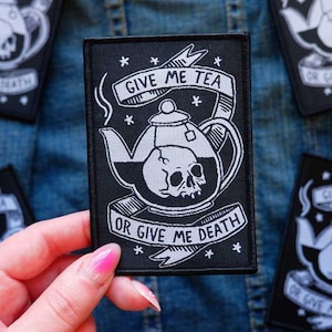 May include: A rectangular black embroidered patch featuring a white line art illustration of a teapot containing a human skull. Above the teapot is a banner with the text GIVE ME TEA and below is a banner with the text OR GIVE ME DEATH.