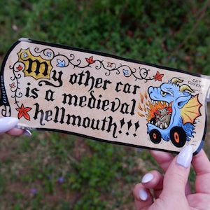 May include: A sticker with the text "my other car is a medieval hellmouth!!!" in a medieval font. The sticker features a blue dragon driving a car with flames and a small creature inside.