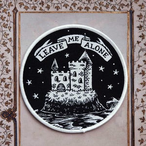 May include: A round black and white patch featuring a castle on a rocky island, surrounded by stars. The banner above reads "LEAVE ME ALONE". The patch has a white border and is set against a decorative background.