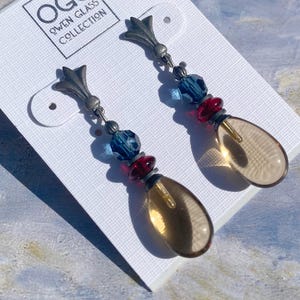 May include: A pair of dangle earrings with a silver-tone metal design. The earrings feature blue and red beads, and teardrop-shaped amber-colored glass pendants. The earrings are displayed on a white card with the text "OGC Owen Glass Collection".
