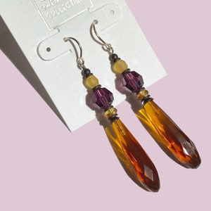 May include: A pair of dangle earrings with amber and purple glass beads. The earrings are on a white card with the text "OGC Owen Glass Collection".