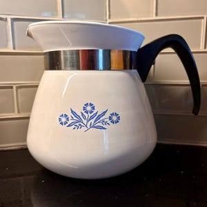 May include: A white ceramic pitcher featuring a blue floral pattern on the front. The pitcher has a wide, rounded body, a metallic silver band encircling the upper section, and a curved black handle extending from the side for easy pouring.
