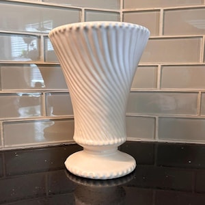 May include: A white ceramic vase featuring a flared, wide opening and a tapered body with a distinct diagonal ribbed texture. The vase stands on a circular pedestal base, with small beaded detailing along the top rim and the base edge.