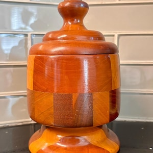 May include: A cylindrical wooden container with a lid, featuring a checkerboard pattern of alternating light and dark brown wood segments. The container has a rounded base and a matching lid with a central knob handle, all with a smooth finish.