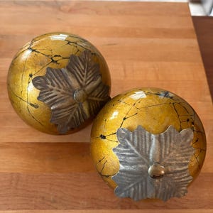 May include: Two spherical decorative objects with a glossy mustard yellow finish featuring a dark black crackle pattern. Each sphere is topped with a metallic silver-toned leaf-shaped ornament that has a textured surface and a central circular stud.